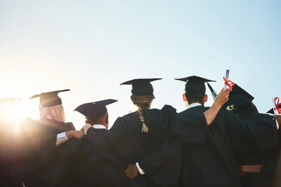 View from back of group of college students in caps and gowns at graduation with bright sunlight in front of them.